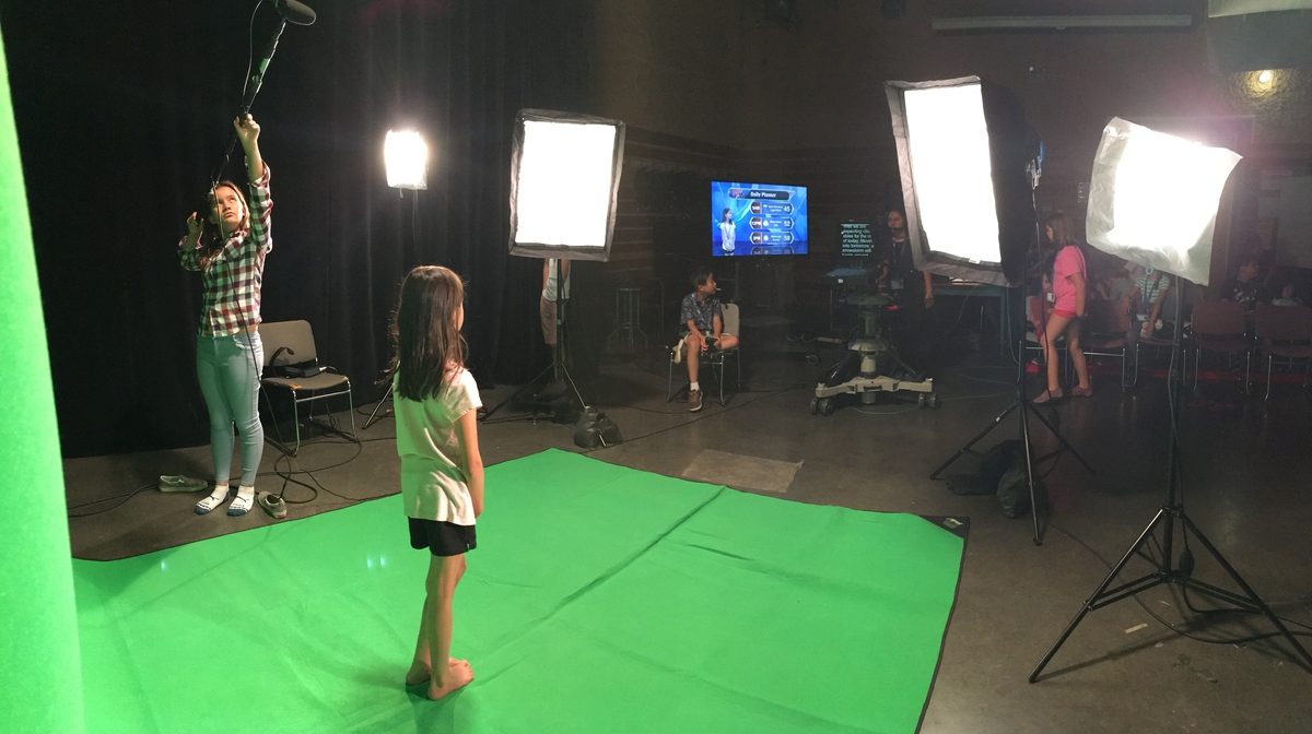 A girl stands on a green screen backdrop at broadcast camp while another girl holds a boom mic