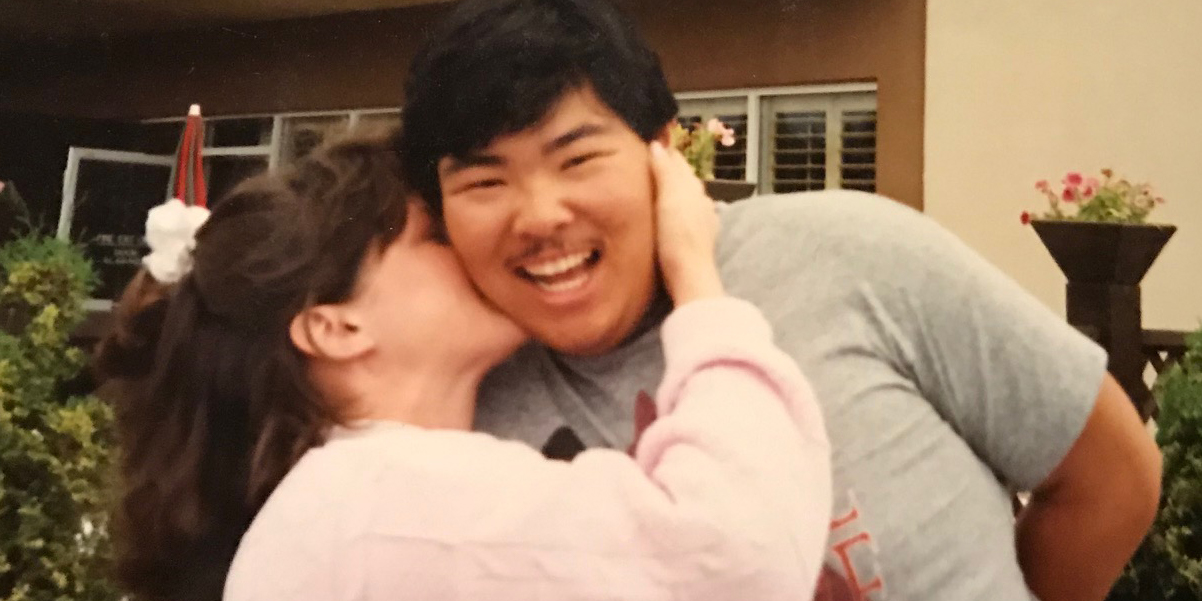 woman in pink sweater kisses a smiling man on the cheek in front of a diner