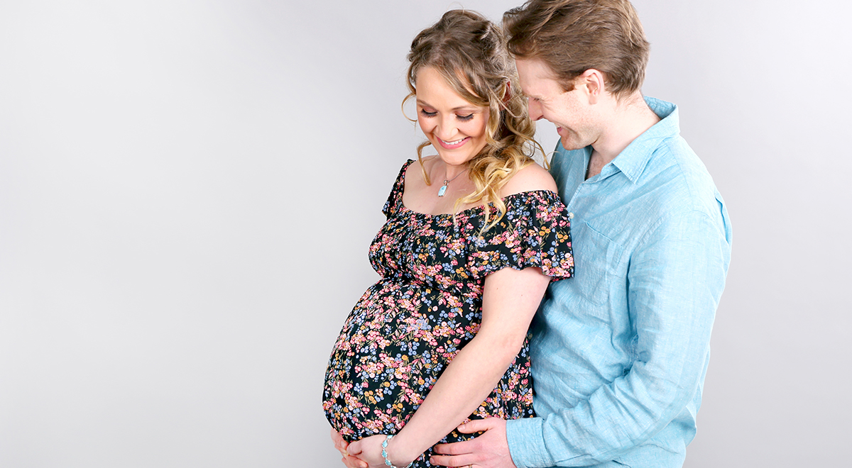 man in blue shirt embraces woman in floral dress, both with a hand on the woman's pregnant belly