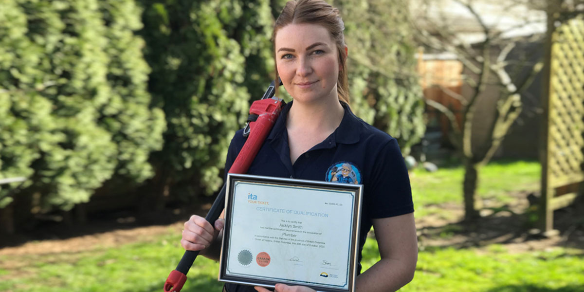 Female plumber standing outside holding a pipe wrench and her Red Seal certification.