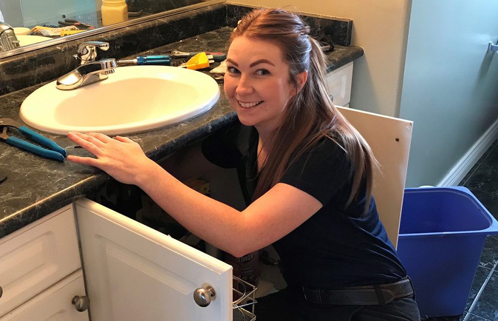 Female plumber kneeling down at a bathroom sink, smiling.
