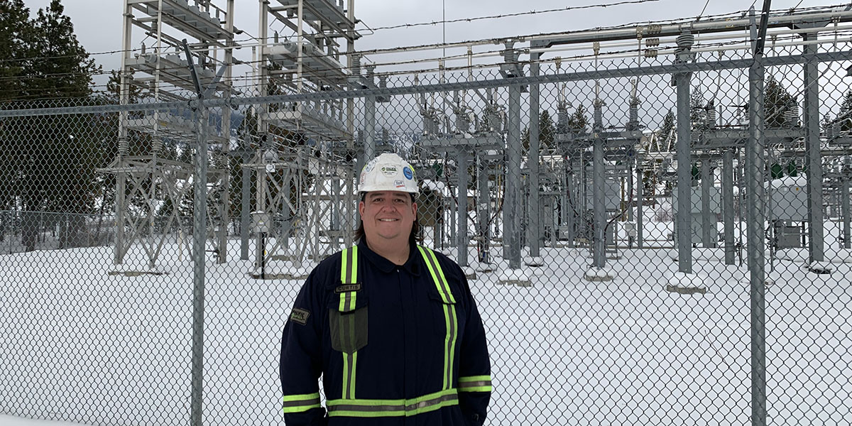 A man wearing safety gear and a hard hat standing in front of a electrical power station smiling.