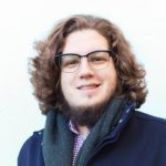 Man with a beard, long curly red-brown hair, navy coat and scarf smiles