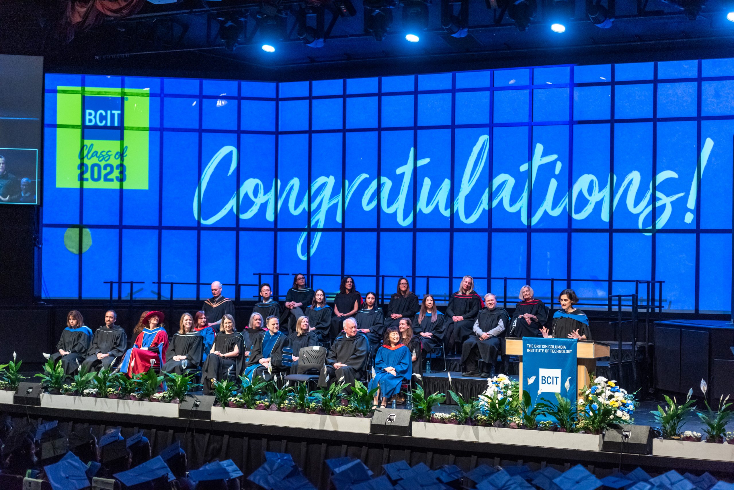 A wide shot of the platform party on the auditorium stage, with a large congratulations text as a background on the digital screen.