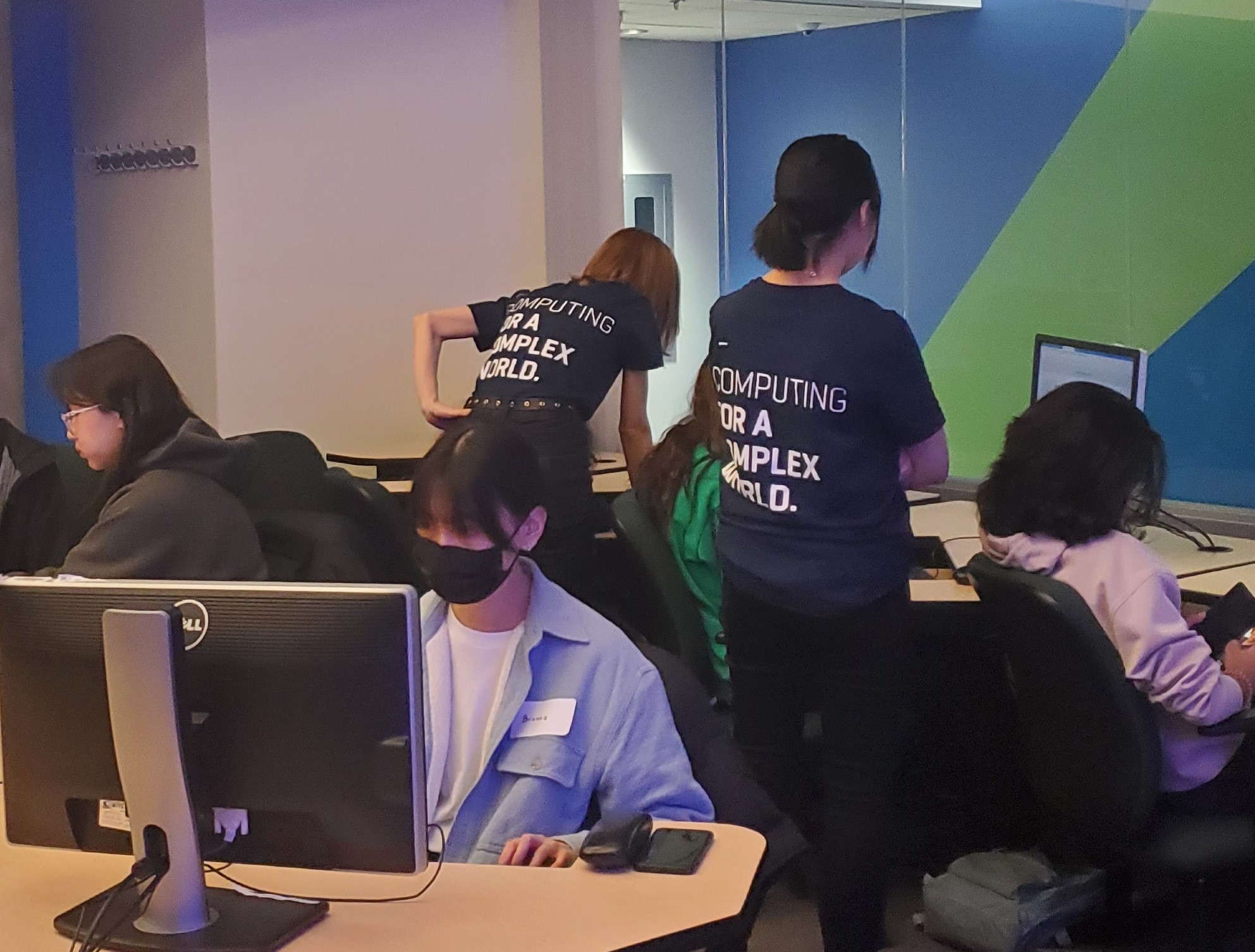 Two young women wearing "Computing for a Complex World" t-shirts help other young women working in a computer lab