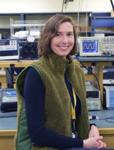 A portrait of Jessica Wilson, BCIT alumna and Assistant Instructor at the BCIT Electrical Engineering program, smiling straight at the camera. She is wearing a dark blue, long-sleeved shirt, with a green vest.