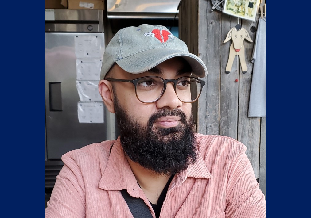 Younger man with blue baseball cap, glasses, and full beard looks into distance. He is wearing a tea rose corduroy shirt and is perhaps in a tech company kitchen