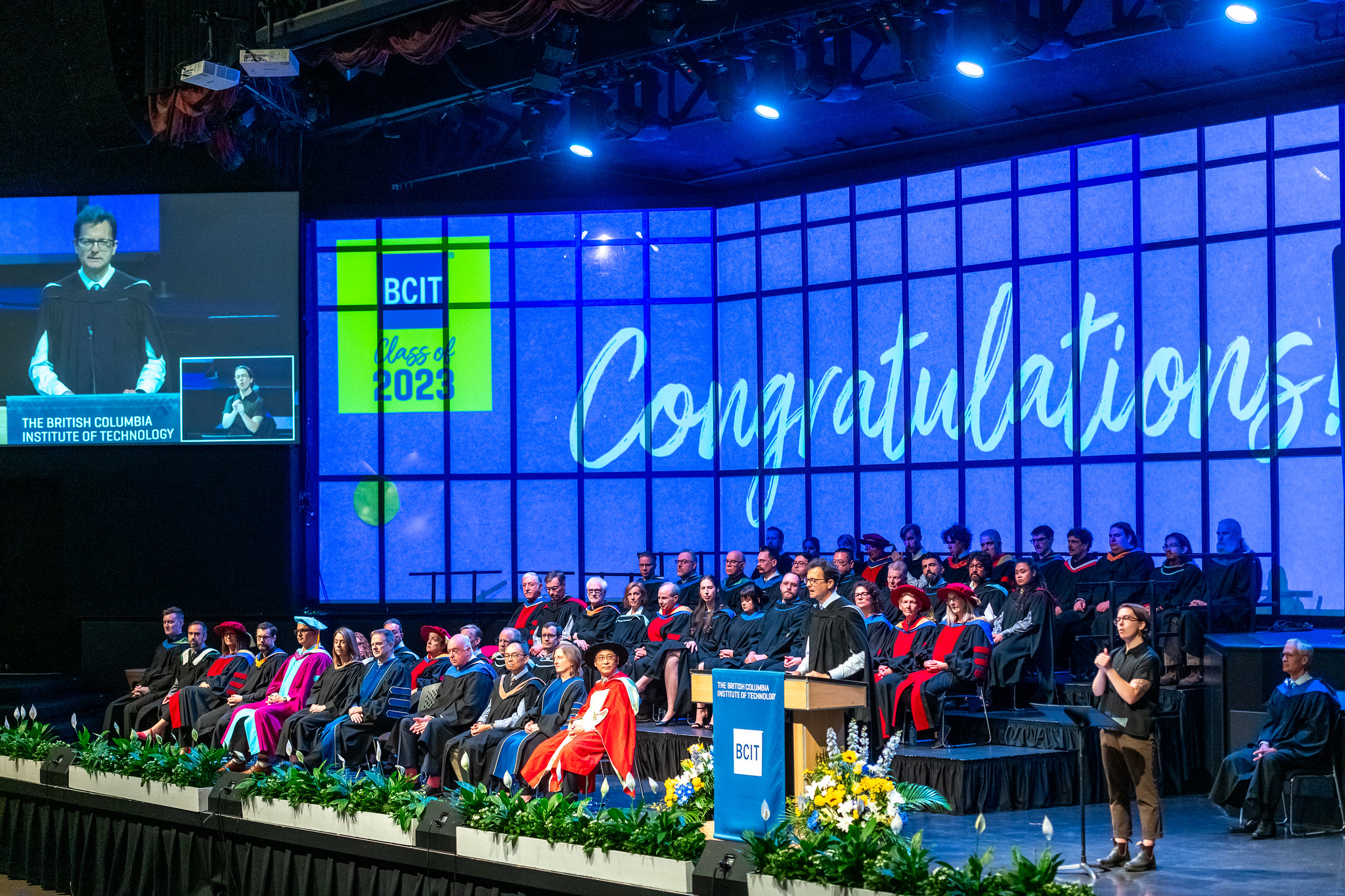 Photo of Willingdon Centre stage with a big screen that says Congratulations Grads of 2023 with people sitting on the stage in grad gowns.