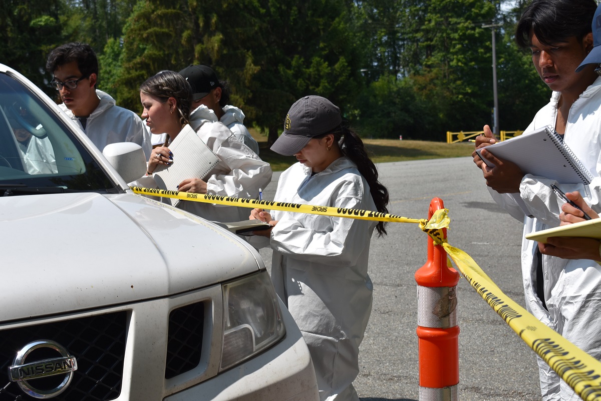 Four students in white full-body hazmat suits take notes at a mock crime scene behind yellow tape. They are looking into a white truck. More students are in the background.