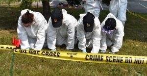 Four young people wearing full-body white hazmat type suits crawl through the grass behind yellow tape that reads "crime scene do not cross"