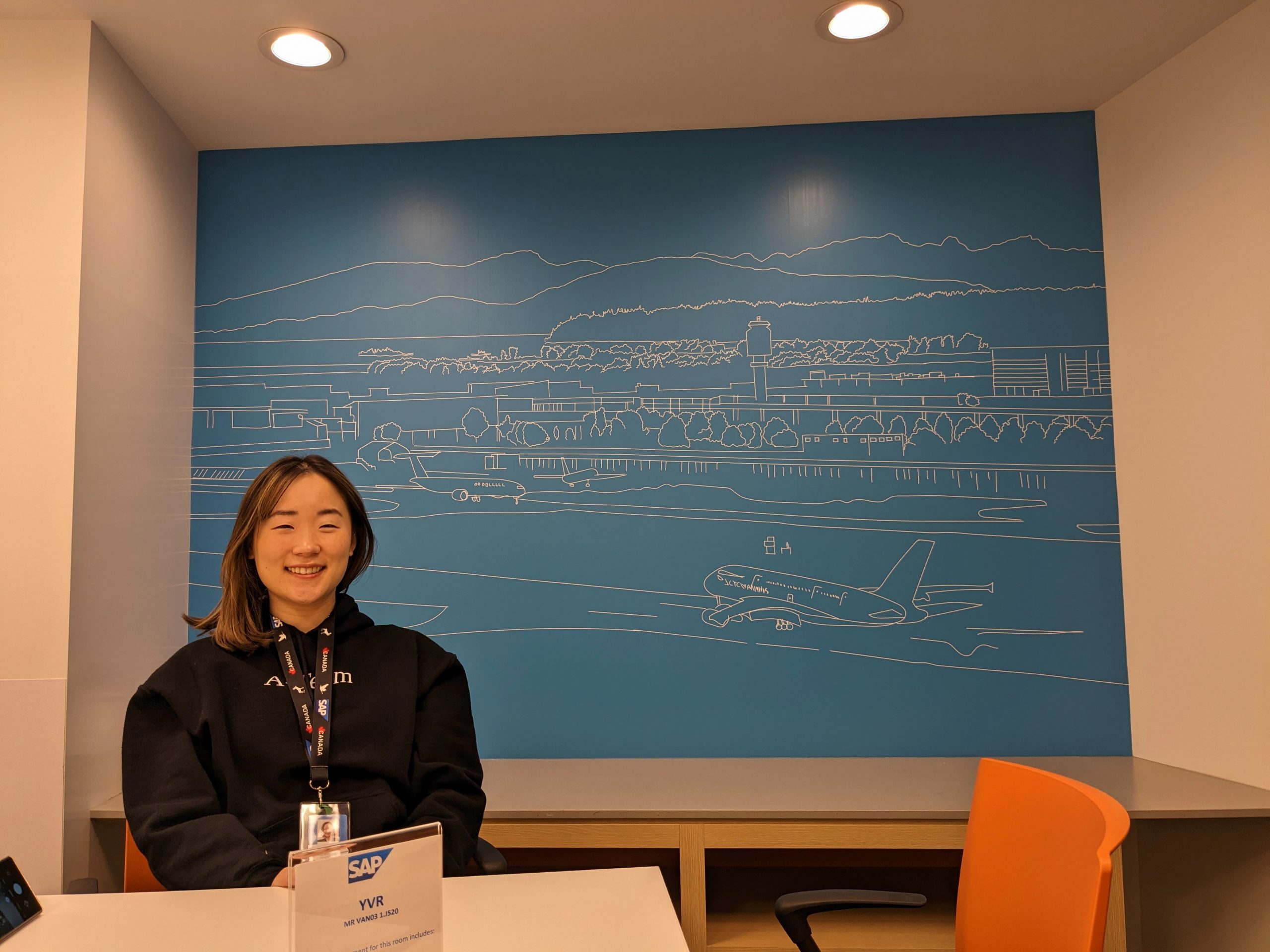 A young woman with midlength dark hair sits in a conference room at SAP. Behind her is a line drawn art piece showing an airport with mountains behind it.