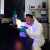 Forensic Investigation instructor Dom Toa wears a full-body white "bunny suit" and glasses in a lab. He is shining a light on a drinking glass. There is a bottle behind him and laboratory shelves with a lab sink and equipment.