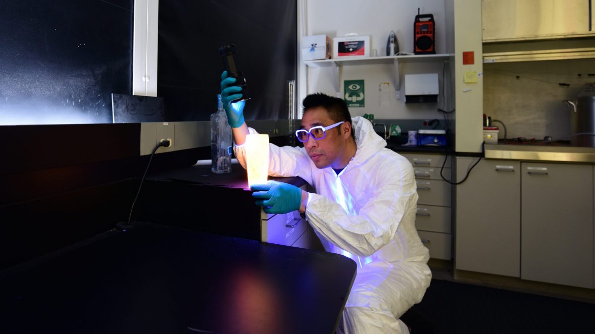 Forensic Investigation instructor Dom Toa wears a full-body white "bunny suit" and glasses in a lab. He is shining a light on a drinking glass. There is a bottle behind him and laboratory shelves with a lab sink and equipment.
