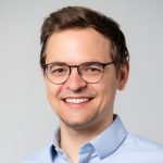 Younger faculty Michal Aibin smiles wearing glasses and a light blue dress shirt