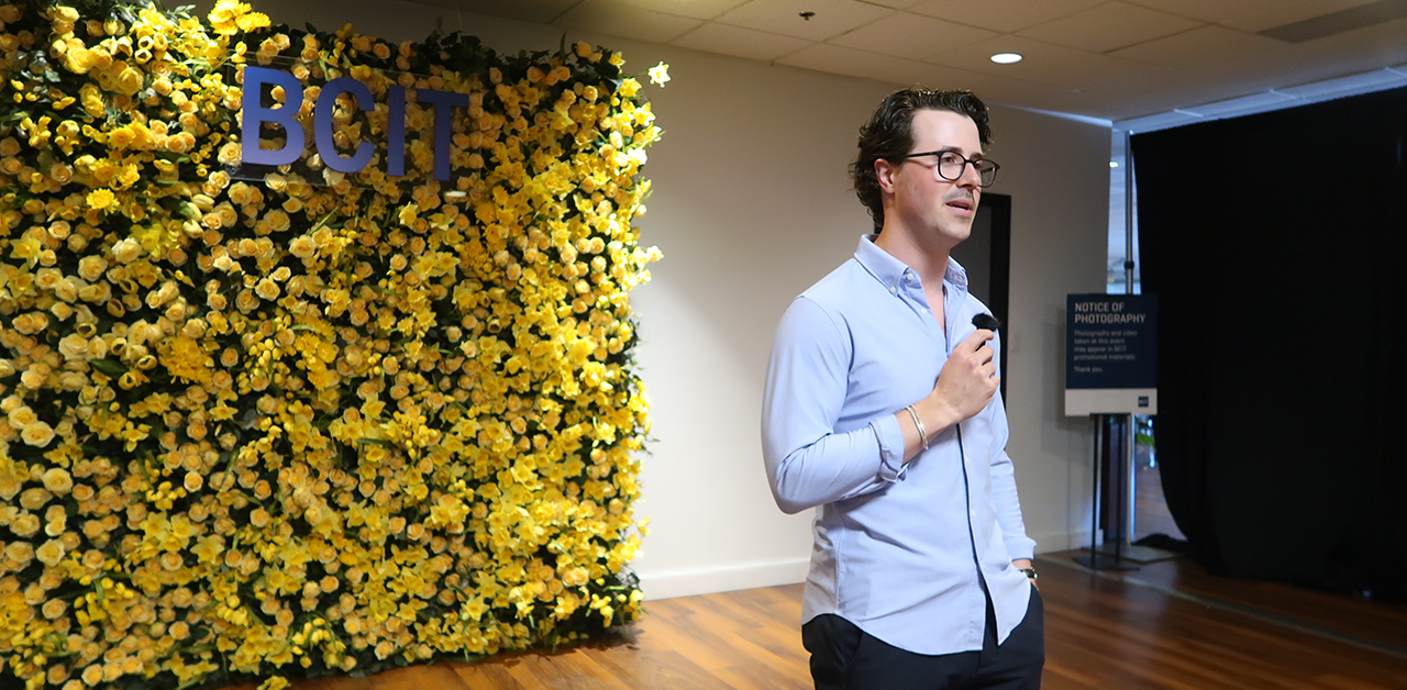 Matt Cox delivers a speech at the BCIT 2024 Spring Convocation Graduating Awards Ceremony. He holds a microphone, and stands in front of a yellow flower wall with a sign on it that reads "BCIT"