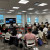 group of mixed demographic adults seated in a circle in a modern meeting room with additional meeting attendees on screen