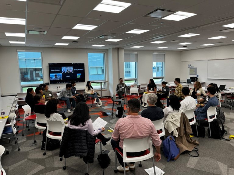 group of mixed demographic adults seated in a circle in a modern meeting room with additional meeting attendees on screen