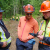 Three people in high vis gear stand in the forest looking at a electronic tablet device. The two people on the right are wearing orange safety helmets, and the person on the left is wearing a navy blue baseball cap. In the background, more forestry workers can be seen wearing high-vis gear and orange safety helmets.