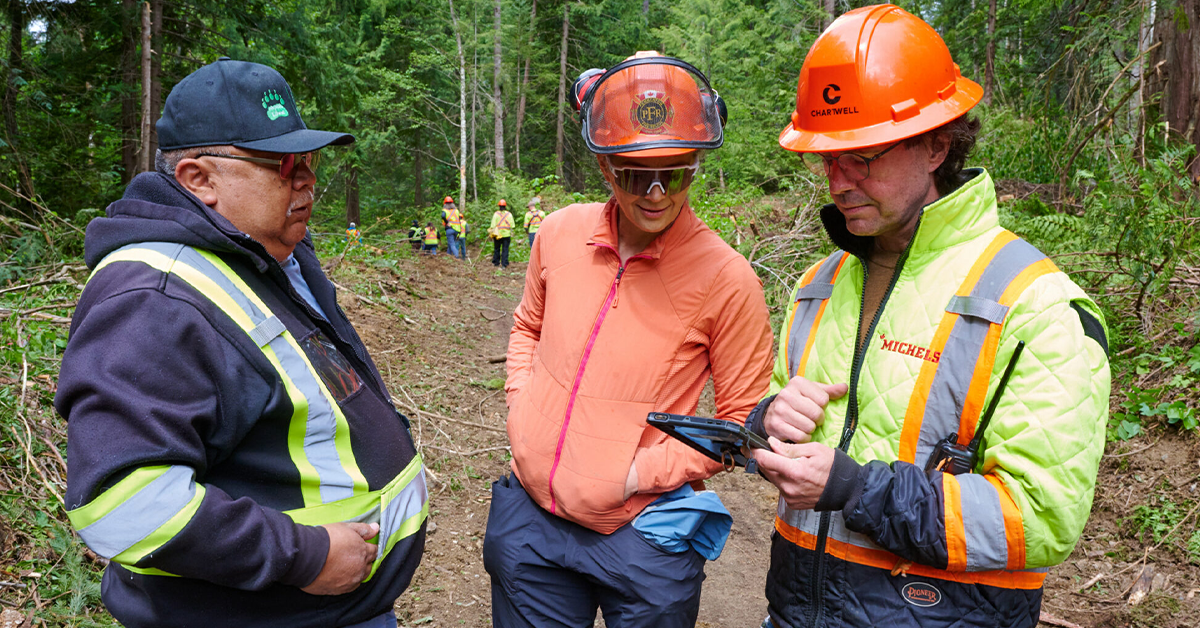 Three people in high vis gear stand in the forest looking at a electronic tablet device. The two people on the right are wearing orange safety helmets, and the person on the left is wearing a navy blue baseball cap. In the background, more forestry workers can be seen wearing high-vis gear and orange safety helmets.